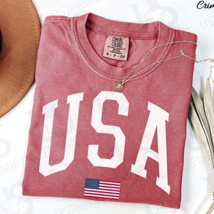 May include: Crimson-coloured t-shirt featuring the word "USA" in large white letters and a small American flag graphic. The shirt has a crew neck and a Comfort Colors tag. A silver necklace with a pendant is draped across the shirt.