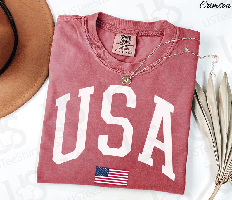 May include: Crimson-coloured t-shirt featuring the word "USA" in large white letters and a small American flag graphic. The shirt has a crew neck and a Comfort Colors tag. A silver necklace with a pendant is draped across the shirt.