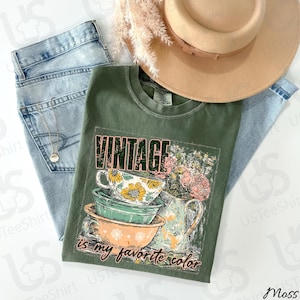 May include: An olive green t-shirt with the word "VINTAGE" printed across the top. The shirt features a graphic of stacked teacups and a floral arrangement. The text "is my favorite color" is printed below the graphic. A light brown hat and blue jeans are also in the image.
