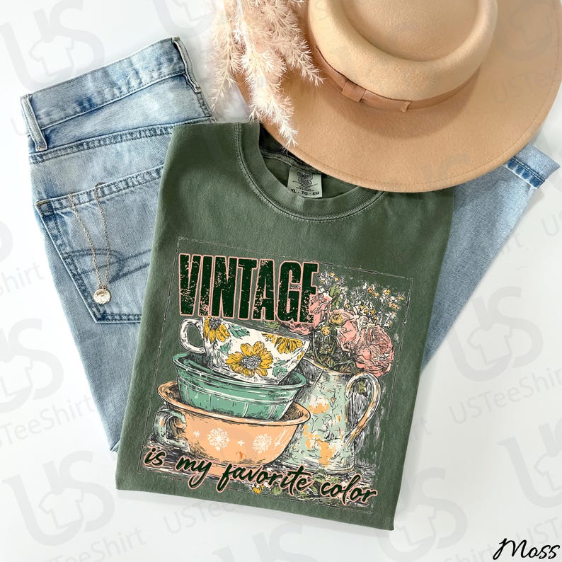 May include: An olive green t-shirt with the word "VINTAGE" printed across the top. The shirt features a graphic of stacked teacups and a floral arrangement. The text "is my favorite color" is printed below the graphic. A light brown hat and blue jeans are also in the image.