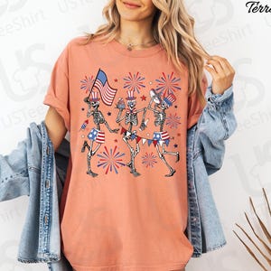 May include: Terracotta-colored t-shirt featuring a festive design of three skeleton figures celebrating with American flags, fireworks, and patriotic attire. The skeletons are depicted in a playful, cartoon style, perfect for Independence Day.