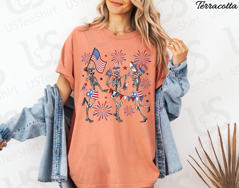 May include: Terracotta-colored t-shirt featuring a festive design of three skeleton figures celebrating with American flags, fireworks, and patriotic attire. The skeletons are depicted in a playful, cartoon style, perfect for Independence Day.