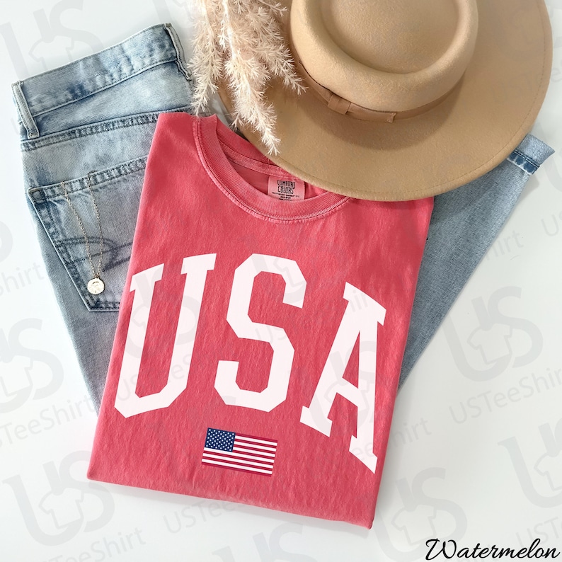 May include: A red t-shirt with the word "USA" in large white letters and a small American flag graphic. The shirt is paired with light-wash denim jeans and a tan hat. The word "Watermelon" is in the bottom right corner.
