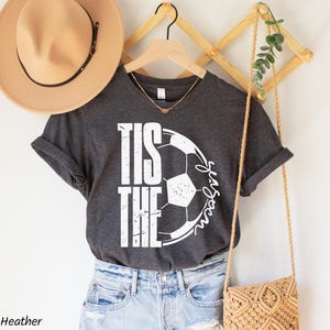 Op de afbeelding: Donkergrijs gem&ecirc;leerd t-shirt met de tekst "Tis the Season" en een voetbalafbeelding. Het shirt hangt aan een houten kleerhanger met een beige hoed en een geweven handtas op de achtergrond.