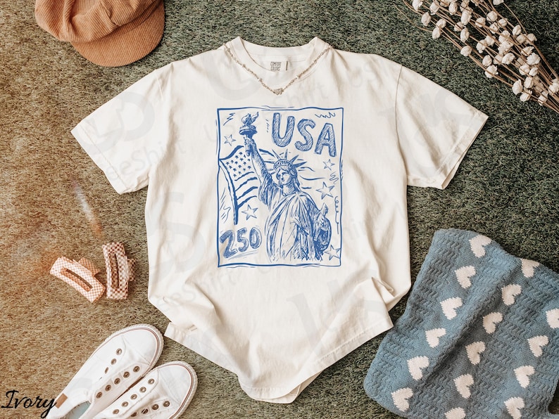 K&ouml;nnte beinhalten: Cremefarbenes T-Shirt mit blauem Aufdruck der Freiheitsstatue, dem Wort "USA" und der Zahl "250". Das Shirt wird mit wei&szlig;en Turnschuhen, einer braunen Kappe und einer blauen Decke mit wei&szlig;en Herzen kombiniert.