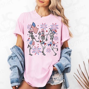 May include: Pink t-shirt featuring a graphic of three skeleton figures celebrating with American flags, hats, and fireworks. The skeletons wear patriotic shorts and wave flags. The shirt has rolled-up sleeves.