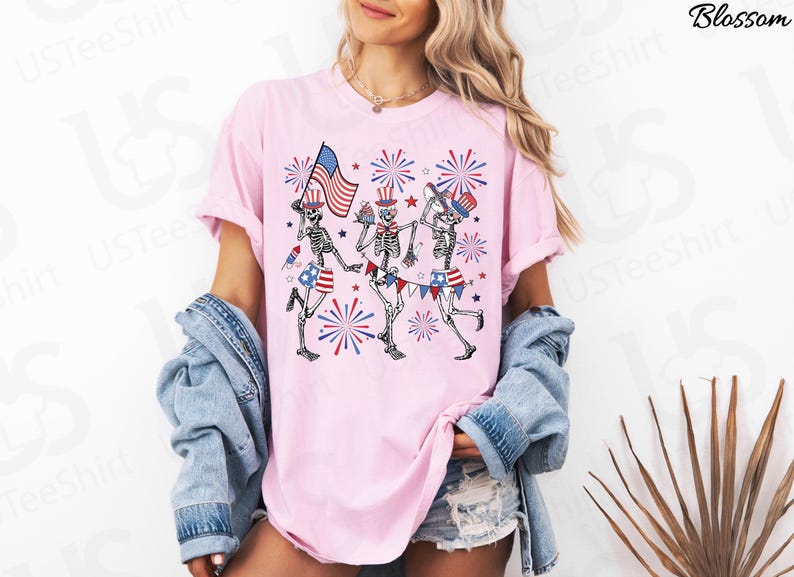 May include: Pink t-shirt featuring a graphic of three skeleton figures celebrating with American flags, hats, and fireworks. The skeletons wear patriotic shorts and wave flags. The shirt has rolled-up sleeves.
