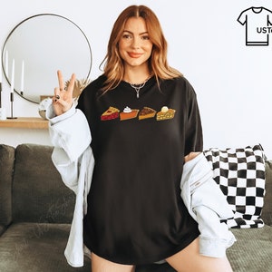 Puede incluir: Una camiseta negra de manga corta con un estampado gr&aacute;fico de cuatro porciones de tarta diferentes dispuestas en una fila horizontal. El dise&ntilde;o incluye una tarta de frutas roja, una tarta de calabaza naranja con nata montada, una tarta de nueces.