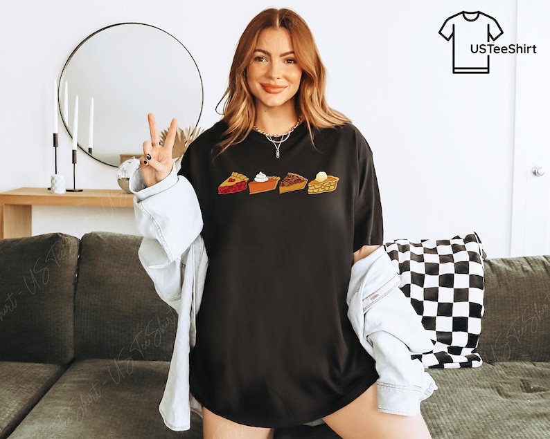 Puede incluir: Una camiseta negra de manga corta con un estampado gr&aacute;fico de cuatro porciones de tarta diferentes dispuestas en una fila horizontal. El dise&ntilde;o incluye una tarta de frutas roja, una tarta de calabaza naranja con nata montada, una tarta de nueces.