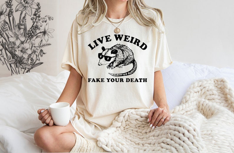 Puede incluir: Camiseta color crema con un gr&aacute;fico negro de una zarig&uuml;eya con gafas de sol y el texto "LIVE WEIRD FAKE YOUR DEATH". La camiseta tiene un corte relajado, adecuada para ropa informal.