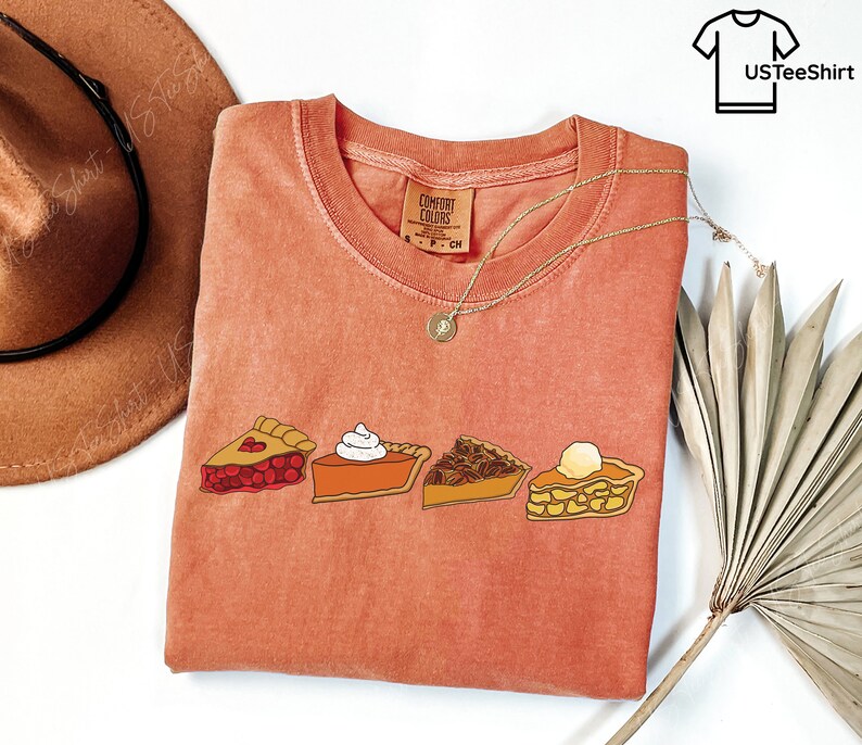 Puede incluir: Una camiseta de manga corta color terracota con un estampado gr&aacute;fico de cuatro porciones de tarta: cereza, calabaza con nata montada, nuez pecana y manzana con una bola de helado. La camiseta tiene cuello redondo y una etiqueta de la marca Comfort Colors.