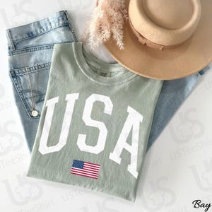 May include: A sage green t-shirt displaying "USA" in white block letters and a small American flag. The shirt is styled with light wash denim jeans, a tan hat, and a gold necklace. The outfit is casual.
