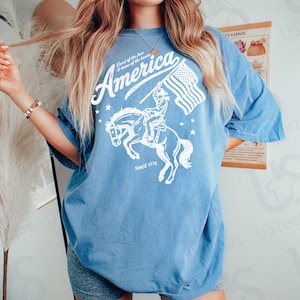 Puede incluir: Camiseta azul claro con un gr&aacute;fico blanco de un jinete a caballo sosteniendo una bandera estadounidense. La palabra "America" est&aacute; encima del gr&aacute;fico, con "Land of the free & home of the brave" y "SINCE 1776" impresos. El texto "Blue Jean" est&aacute; en la esquina superior derecha.