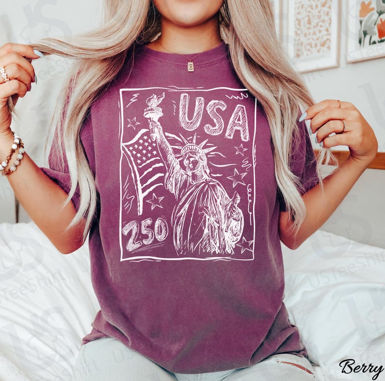 K&ouml;nnte beinhalten: Beerenfarbenes T-Shirt mit einer wei&szlig;en Skizze der Freiheitsstatue, der amerikanischen Flagge und dem Wort "USA". Die Zahl "250" ist ebenfalls im Design enthalten. Das Shirt ist ein bequemer, l&auml;ssiger Stil.