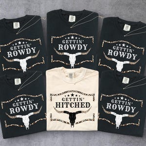 Op de afbeelding: Een verzameling zwarte en crèmekleurige t-shirts. De zwarte shirts hebben de tekst "GETTIN' ROWDY" boven een afbeelding van een stierschedel. Eén crèmekleurig shirt zegt "GETTIN' HITCHED" met een vergelijkbaar ontwerp. De shirts zijn opgevouwen.