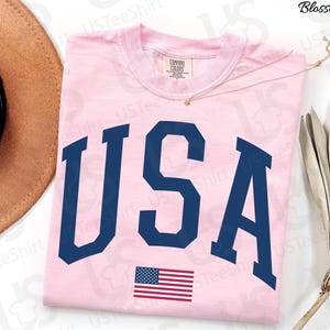 May include: A pale pink t-shirt featuring the word "USA" in navy blue capital letters and a small American flag. The shirt is made of a soft material. The design is patriotic. The t-shirt is likely a comfort fit.