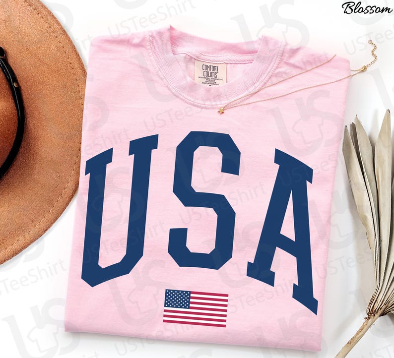May include: A pale pink t-shirt featuring the word "USA" in navy blue capital letters and a small American flag. The shirt is made of a soft material. The design is patriotic. The t-shirt is likely a comfort fit.