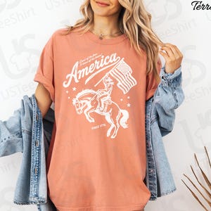 Puede incluir: Camiseta color terracota con un gr&aacute;fico blanco que representa a un jinete a caballo sosteniendo una bandera estadounidense. El texto "America" y "Land of the free, Home of the brave" est&aacute;n sobre el jinete, con "Since 1776" debajo.