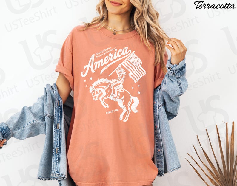 Puede incluir: Camiseta color terracota con un gr&aacute;fico blanco que representa a un jinete a caballo sosteniendo una bandera estadounidense. El texto "America" y "Land of the free, Home of the brave" est&aacute;n sobre el jinete, con "Since 1776" debajo.