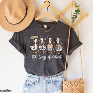 May include: Dark grey heather t-shirt featuring a whimsical design of four cartoon ducks, each holding school supplies and wearing different outfits. The text "100 Days of School" is printed below the ducks. A tan hat and purse are also visible.