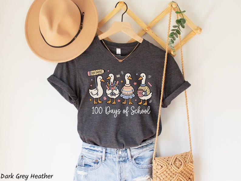 May include: Dark grey heather t-shirt featuring a whimsical design of four cartoon ducks, each holding school supplies and wearing different outfits. The text "100 Days of School" is printed below the ducks. A tan hat and purse are also visible.