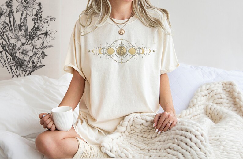 May include: Cream-colored t-shirt with a celestial design featuring suns, moons, and geometric shapes. The shirt has a relaxed fit and is made of soft material. The design is in gold and beige tones. The shirt is part of a clothing collection.