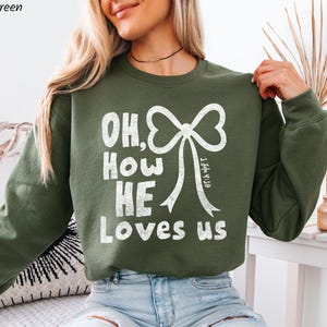 May include: Military green sweatshirt with the text "OH, How HE Loves us" in white, featuring a bow graphic. The sweatshirt is a long-sleeved crew neck style.