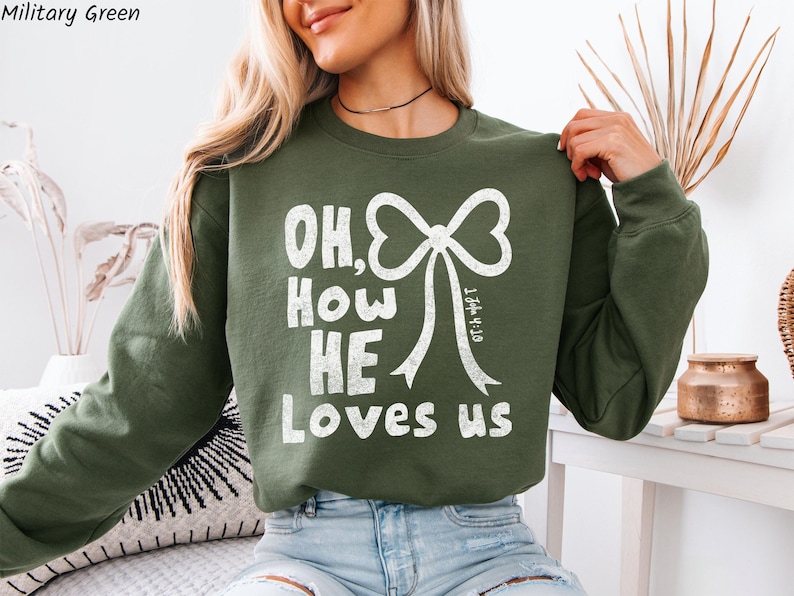 May include: Military green sweatshirt with the text "OH, How HE Loves us" in white, featuring a bow graphic. The sweatshirt is a long-sleeved crew neck style.