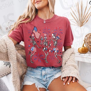 May include: Crimson-colored t-shirt featuring a festive design of dancing skeletons celebrating Independence Day. The skeletons are adorned with patriotic attire, including top hats, flags, and star-spangled shorts. Fireworks and the American flag complete the design.