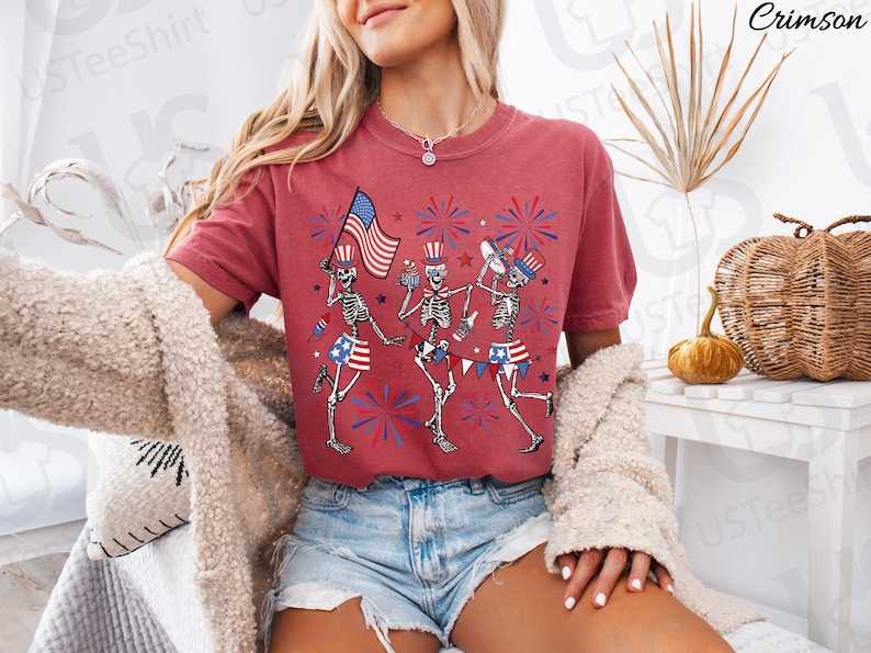 May include: Crimson-colored t-shirt featuring a festive design of dancing skeletons celebrating Independence Day. The skeletons are adorned with patriotic attire, including top hats, flags, and star-spangled shorts. Fireworks and the American flag complete the design.