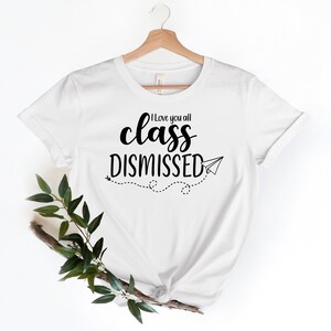 May include: White t-shirt with the text "I love you all class DISMISSED" in black script and a paper airplane graphic. The shirt is hanging on a wooden hanger with a sprig of greenery in the lower left corner.