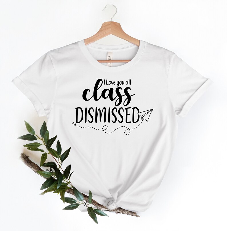 May include: White t-shirt with the text "I love you all class DISMISSED" in black script and a paper airplane graphic. The shirt is hanging on a wooden hanger with a sprig of greenery in the lower left corner.