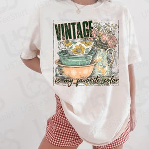 May include: Ivory-colored t-shirt featuring a vintage-style graphic. The graphic includes stacked teacups and bowls, a floral pitcher, and the text "VINTAGE is my favorite color." The shirt is paired with red and white checkered shorts.