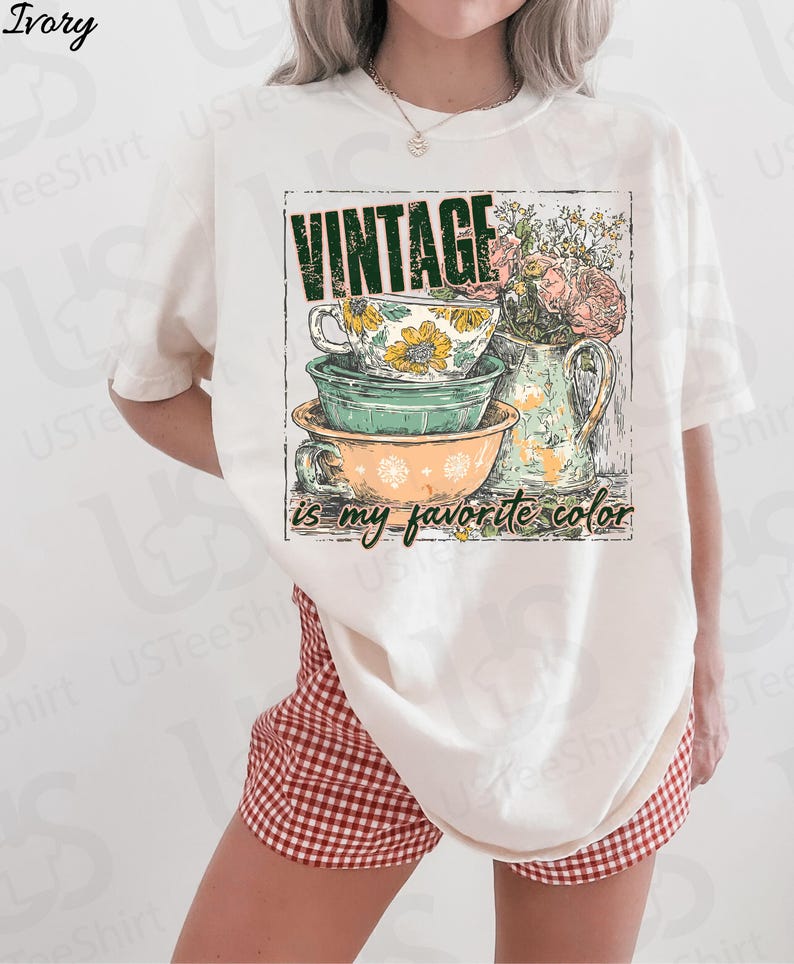 May include: Ivory-colored t-shirt featuring a vintage-style graphic. The graphic includes stacked teacups and bowls, a floral pitcher, and the text "VINTAGE is my favorite color." The shirt is paired with red and white checkered shorts.