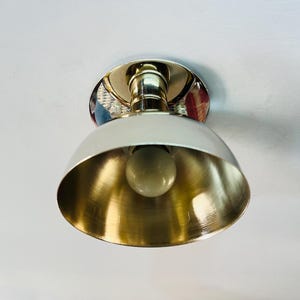 May include: A ceiling-mounted light fixture with a white, dome-shaped shade and a gold-colored interior. The fixture has a polished chrome base and a visible light bulb. The design is simple and modern, suitable for various interior styles.
