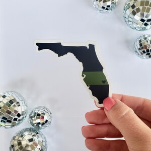 May include: A black and green sticker of the state of Florida with a small heart in the center of the green stripe. The sticker is being held by a hand.