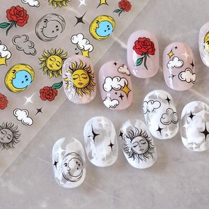 May include: Nail art stickers featuring celestial designs. The sheet includes suns, moons, stars, clouds, and roses. Some nails are painted with the designs, while others have a clear base with the stickers applied. The color palette includes yellow, red, white, and black.