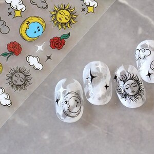K&ouml;nnte beinhalten: Nagelkunst-Aufkleber mit himmlischen Designs. Das Blatt enth&auml;lt Monde, Sonnen, Wolken, Sterne und Rosen. Die N&auml;gel sind wei&szlig; lackiert mit schwarzen himmlischen Designs, darunter Monde, Sonnen, Wolken und Sterne.