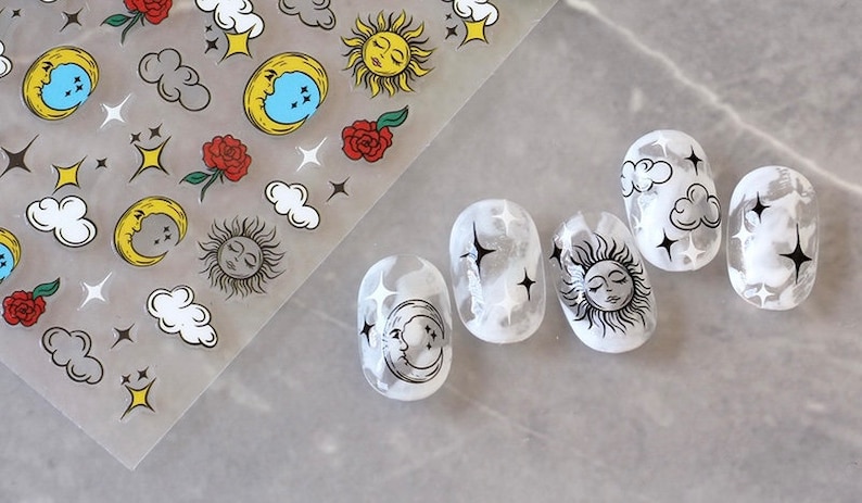 K&ouml;nnte beinhalten: Nagelkunst-Aufkleber mit himmlischen Designs. Das Blatt enth&auml;lt Monde, Sonnen, Wolken, Sterne und Rosen. Die N&auml;gel sind wei&szlig; lackiert mit schwarzen himmlischen Designs, darunter Monde, Sonnen, Wolken und Sterne.