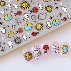 K&ouml;nnte beinhalten: Nagelkunst-Aufkleber mit himmlischen und floralen Designs. Die Aufkleber zeigen Sonnen, Monde, Wolken, Sterne und Rosen in Gelb, Rot und Wei&szlig;. Mehrere N&auml;gel sind mit den Aufklebern verziert, um die Designs zu pr&auml;sentieren.
