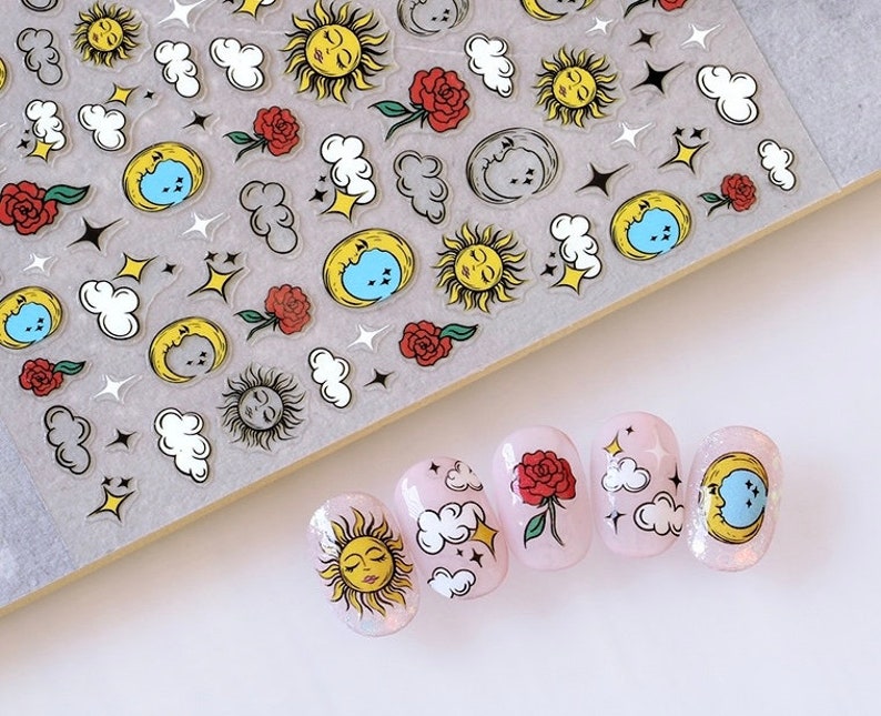 K&ouml;nnte beinhalten: Nagelkunst-Aufkleber mit himmlischen und floralen Designs. Die Aufkleber zeigen Sonnen, Monde, Wolken, Sterne und Rosen in Gelb, Rot und Wei&szlig;. Mehrere N&auml;gel sind mit den Aufklebern verziert, um die Designs zu pr&auml;sentieren.