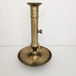 May include: A vintage brass candlestick holder with a round base and a tall, cylindrical stem. The candlestick has a small knob on the side and a flared top to hold a candle. The brass has a warm, aged patina.