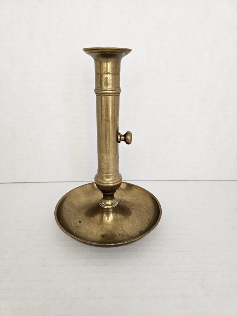 May include: A vintage brass candlestick holder with a round base and a tall, cylindrical stem. The candlestick has a small knob on the side and a flared top to hold a candle. The brass has a warm, aged patina.