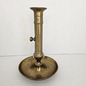 May include: A vintage brass candlestick holder with a round base and a tall, cylindrical stem. The candlestick has a small knob on the side and a flared top to hold a candle. The brass has a warm, aged patina.