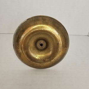 May include: A close-up view of the underside of a brass object. The object has a circular base with a slightly concave surface and a small hole in the center. The brass has a warm, golden hue with some signs of wear.