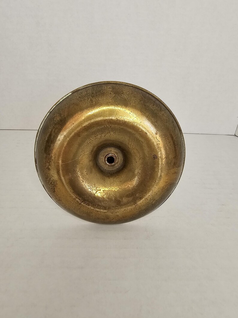 May include: A close-up view of the underside of a brass object. The object has a circular base with a slightly concave surface and a small hole in the center. The brass has a warm, golden hue with some signs of wear.