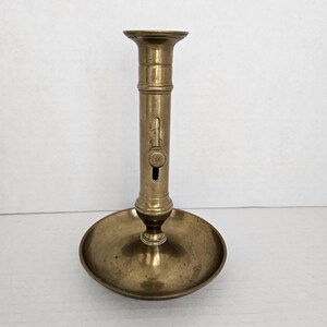 May include: A vintage brass candlestick holder with a round base and a tall, cylindrical stem. The candlestick has a small, circular opening and a mechanism on the side. The overall design is simple and elegant.