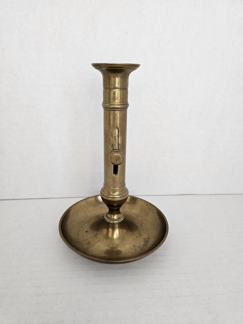 May include: A vintage brass candlestick holder with a round base and a tall, cylindrical stem. The candlestick has a small, circular opening and a mechanism on the side. The overall design is simple and elegant.