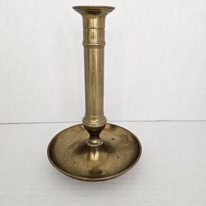 May include: A vintage brass candlestick holder with a round base and a tall, cylindrical stem. The candlestick has a slightly flared top to hold a candle. The brass has a warm, aged patina, suggesting its antique nature. The design is simple and elegant.