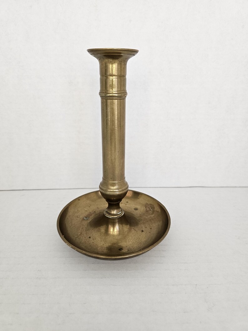 May include: A vintage brass candlestick holder with a round base and a tall, cylindrical stem. The candlestick has a slightly flared top to hold a candle. The brass has a warm, aged patina, suggesting its antique nature. The design is simple and elegant.